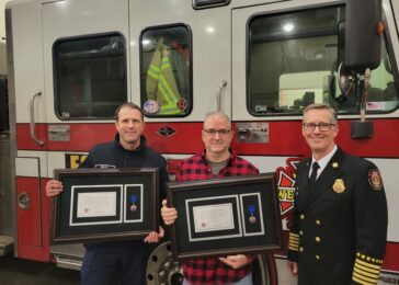 Three firefighters standing with merit awards in front of a fire engine