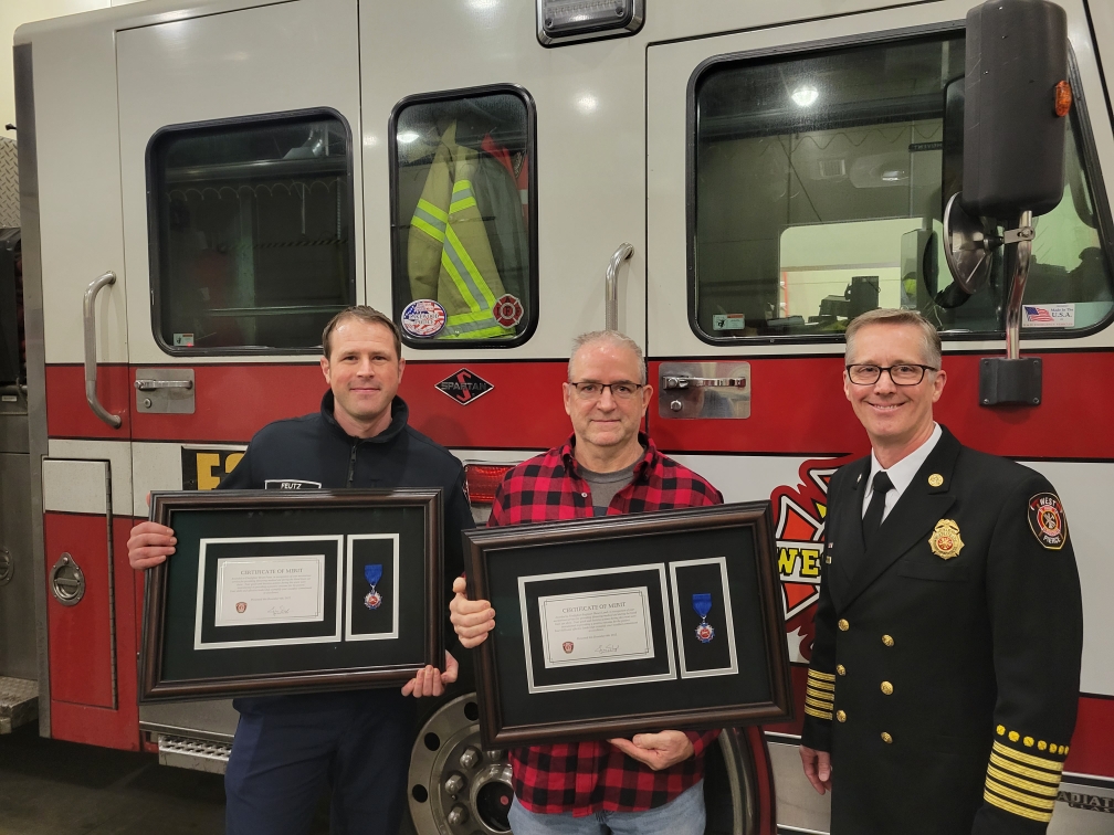 Three firefighters standing with merit awards in front of a fire engine